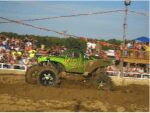 A green monster truck performing in a muddy arena with a crowd watching.